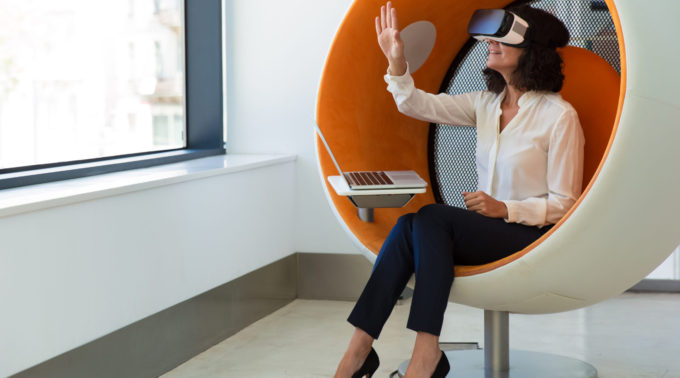 Businesswoman playing virtual game in studio. Woman in office clothes and virtual reality glasses sitting in interactive chair with laptop and touching air. VR game concept